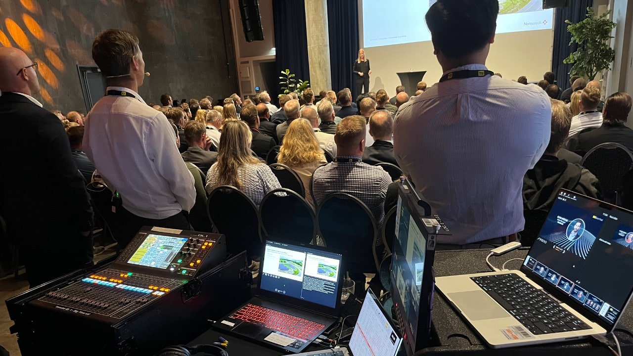 A conference presentation in a large auditorium with a large audience. In the foreground, audio-visual equipment, including a control panel and a laptop, captures the event. A speaker is visible on stage.