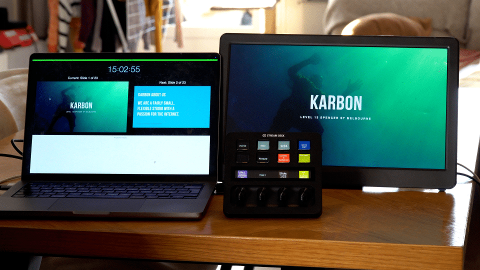 A laptop and a monitor set up on a wooden table. The laptop displays a timer and text labeled "KARBON," while the monitor shows a centered graphic with the same label. A control panel with buttons is placed in front of the monitor.