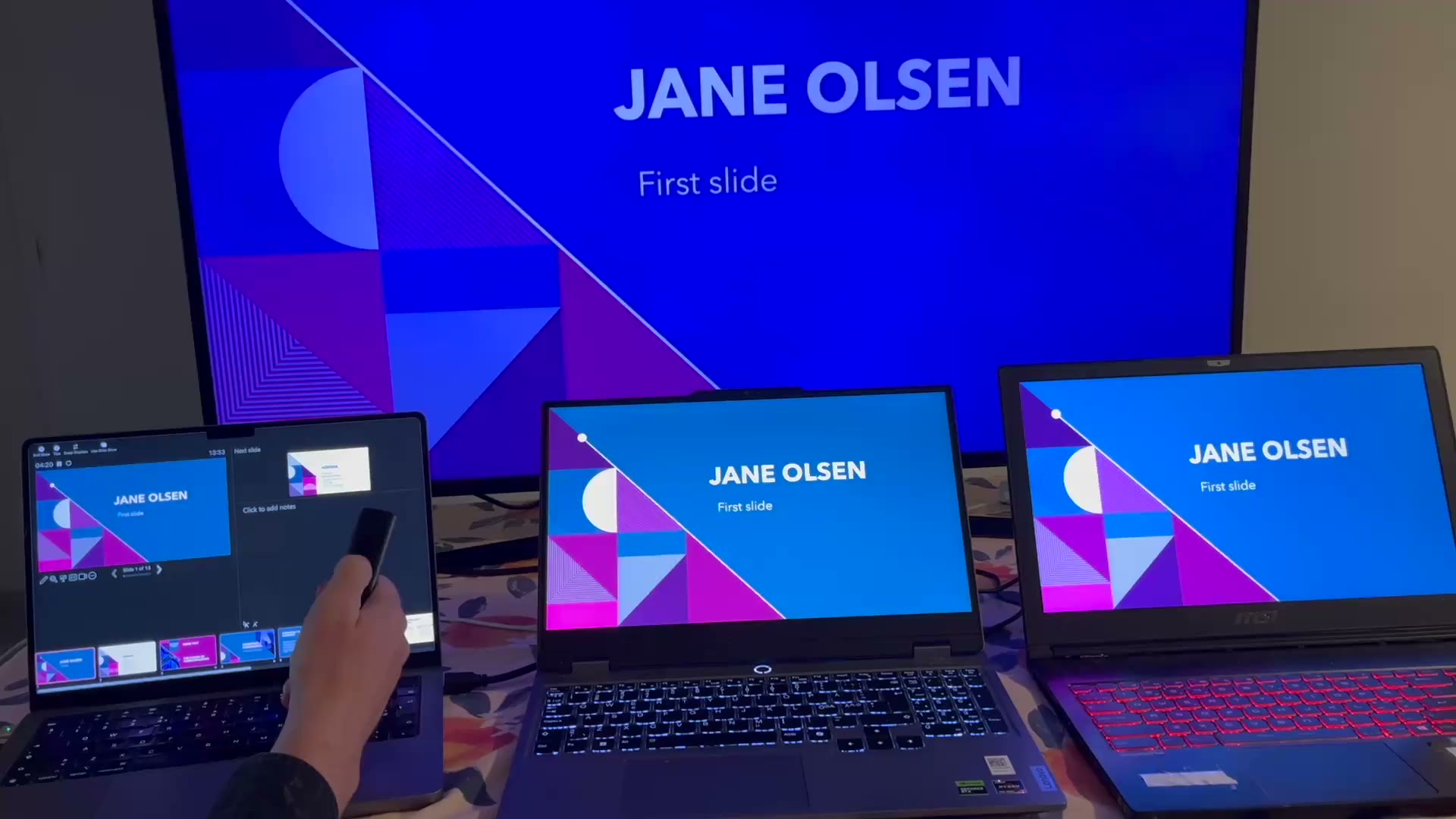 A Mac and two Windows laptops on a desk, all showing the same slide of a presentation, with a presentation clicker in the foreground.